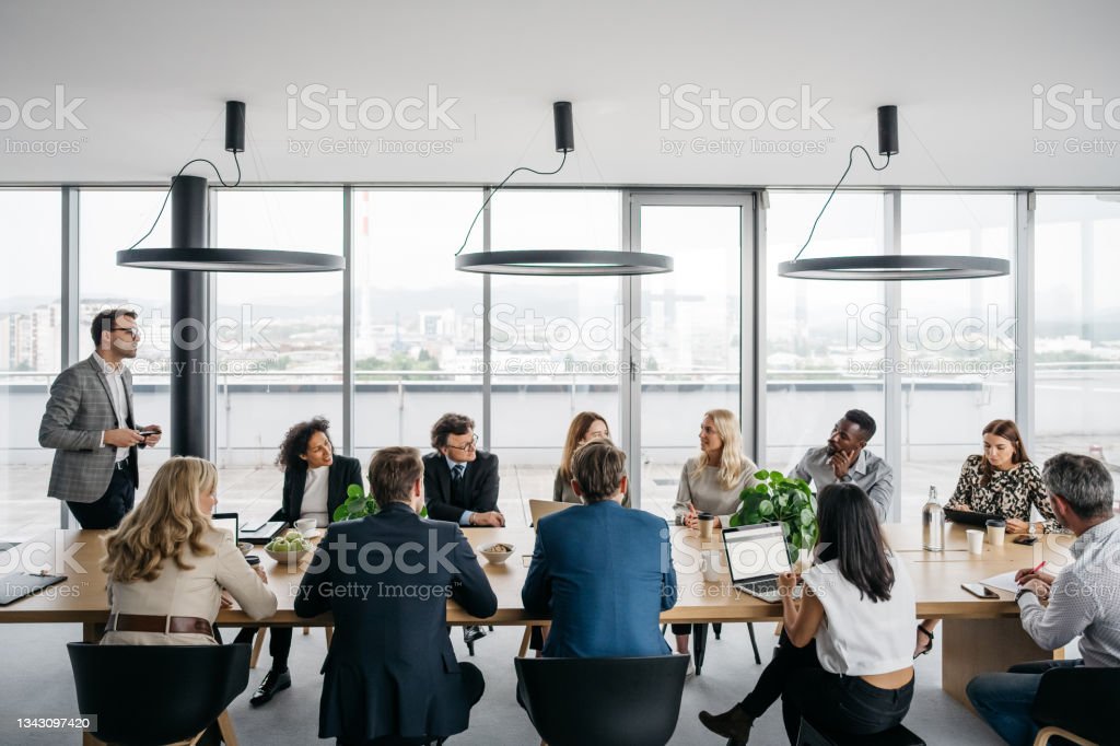Una foto di una riunione d'affari in un ufficio moderno con grandi finestre. Un uomo d'affari è in piedi mentre i suoi colleghi sono seduti. Sono vestiti elegantemente. Foto orizzontale in interni alla luce del giorno.
