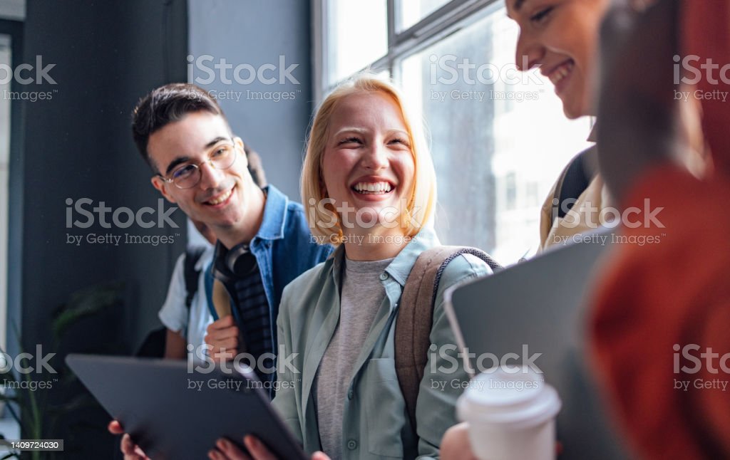 Sorridente gruppo multietnico di amici in fila e appoggiati a una finestra mentre utilizzano un tablet digitale, parlano e ridono insieme.